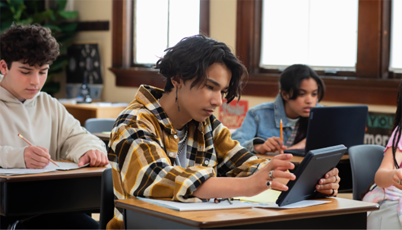alunos em uma sala de aula. um garoto a frente utilizando um tablet, outro gatoro atrás escrevendo em uma folha e uma menina ao fundo com um notebook.