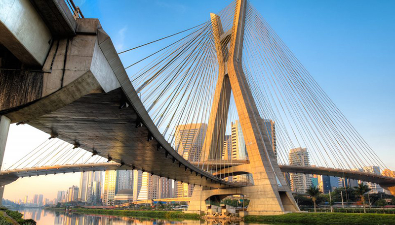 Grande ponte com cabos de aço inclinados, cruzando um rio e conectando duas áreas da cidade de São Paulo.
