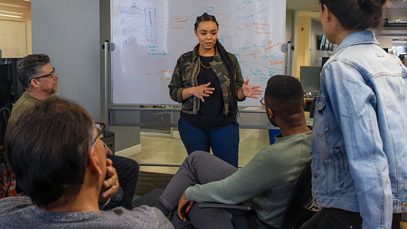 Desenvolvedora negra falando em frente a um quadro branco durante uma reunião de equipe.