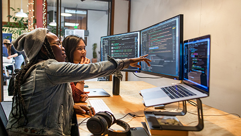 Pessoas reais, escritórios reais. Duas desenvolvedoras trabalhando e colaborando em um escritório empresarial. Elas estão usando Visual Studio em um MacBook Pro com um conjunto de monitores múltiplos.