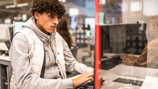 Um aluno está sentado em frente ao computador de uma biblioteca ou sala de aula, digitando no teclado. Fileiras de outras estações de computador são visíveis em segundo plano.