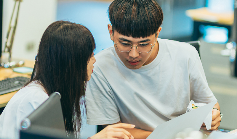 Duas pessoas colaboram em um projeto em uma mesa de escritório. Eles estão olhando a papelada e parecem estar discutindo algo