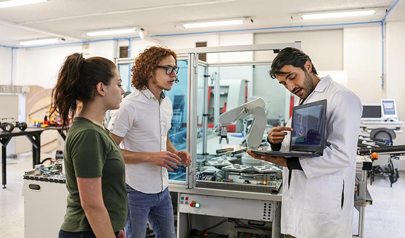 um homem segurando um notebook enquanto apresenta para outro homem e uma mulher em um laboratório com um braço robótico ao fundo