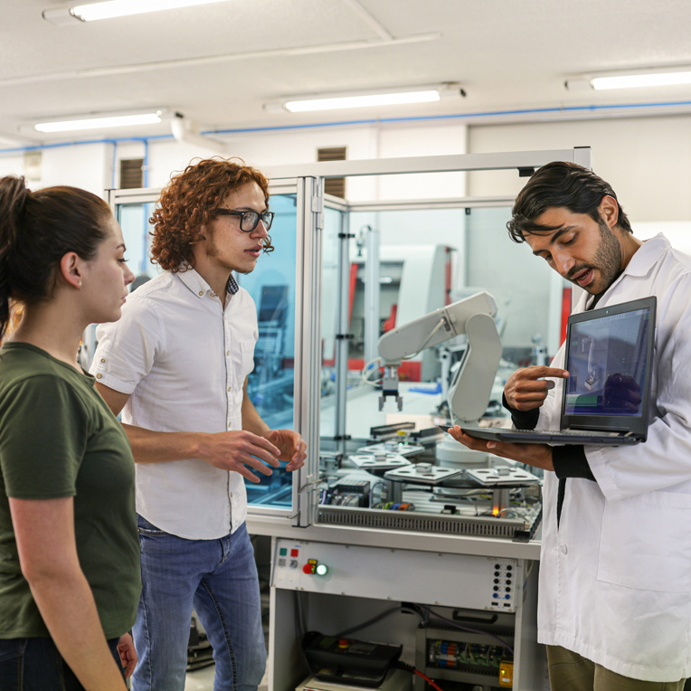 um homem segurando um notebook enquanto apresenta para outro homem e uma mulher em um laboratório com um braço robótico ao fundo