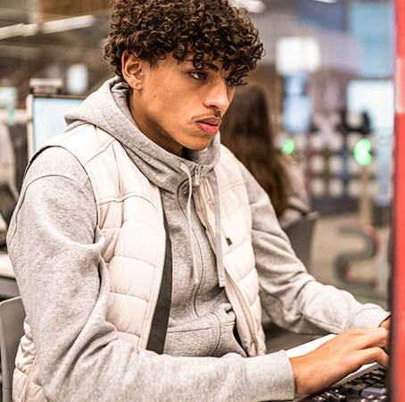 Um aluno está sentado em frente ao computador de uma biblioteca ou sala de aula, digitando no teclado. Fileiras de outras estações de computador são visíveis em segundo plano.