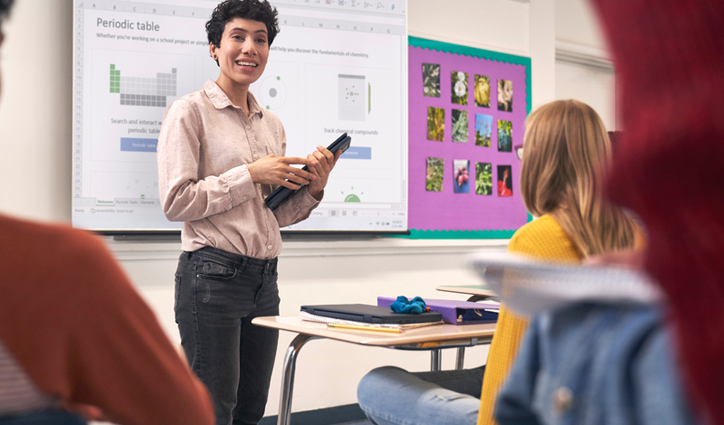 uma professora mostrando algo para os alunos em um notebook sobre a mesa. Ao fundo uma lousa com a palavra SCIENCE escrita