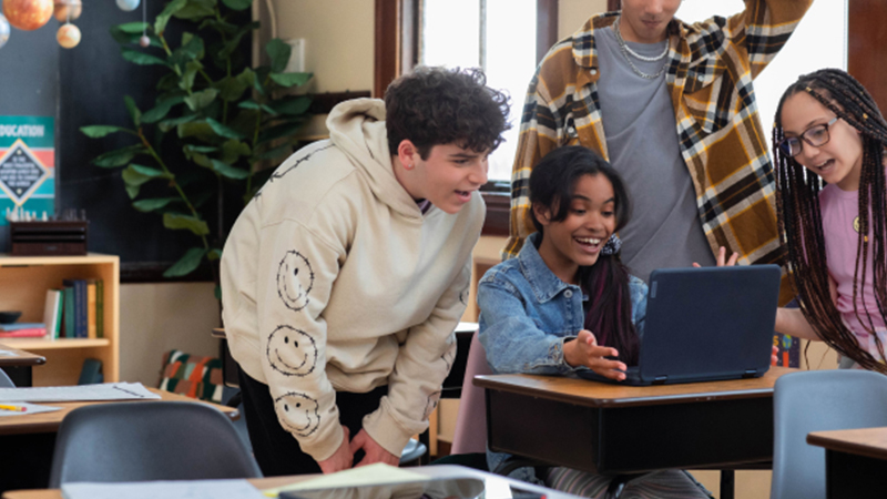 Um grupo de estudantes masculinos e femininos em uma sala de aula escolar está reunido em torno de um laptop aberto em uma mesa e conversando.