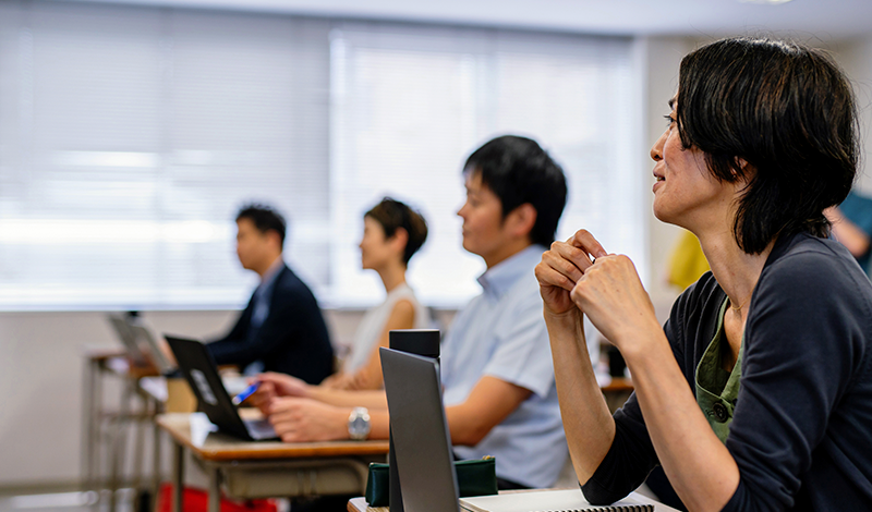 Uma mulher na sala de aula escuta atentamente, com as mãos entrelaçadas. Vários outros estudantes estão sentados em carteiras com laptops, um pouco fora de foco. Grandes janelas proporcionam luz natural.