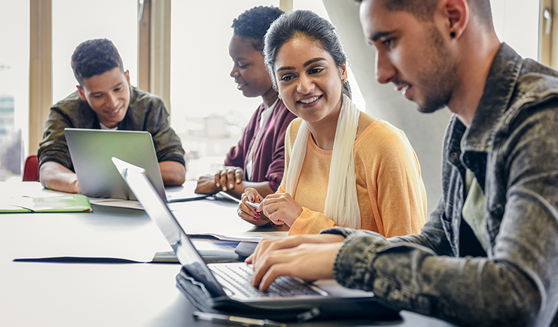 Estudantes universitários colaboram em um projeto, trabalhando juntos em uma mesa com laptops.